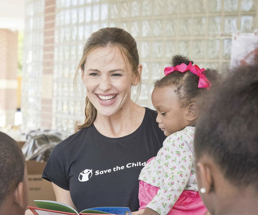 Jennifer Garner holding a child and reading a book, wearing a 'Save the Children' shirt.